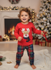 A toddler stands barefoot in a festive holiday setting, wearing a red long-sleeve Christmas pajama top with a reindeer face graphic and matching plaid pajama pants in red, green, and blue. The background features a decorated Christmas tree, a glowing fireplace, and wrapped gifts.
