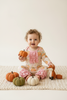 A toddler girl sits cross-legged on a cream knit blanket, wearing a long-sleeve pajama top and matching ruffled pants in a fall-themed floral print. She holds a small orange pumpkin, with other decorative pumpkins and a plush fox toy around her against a clean off-white background.