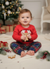 A baby sitting cross-legged on a cream blanket in a holiday-themed room, wearing a red long-sleeve Christmas shirt with a white reindeer graphic and red, green, and blue plaid pants. The background features a decorated Christmas tree, wrapped gifts, and festive greenery.