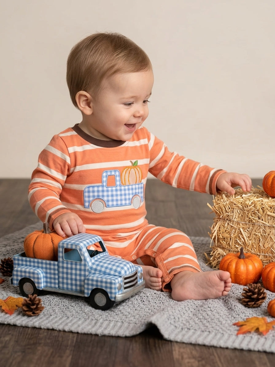 A baby sitting on a knit blanket wearing an orange and white striped long-sleeve pajama with a blue-and-white check truck and pumpkin graphic. The baby is surrounded by autumn props including pumpkins, pinecones, a toy truck, and a hay bale.