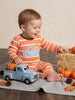 A baby sitting on a knit blanket wearing an orange and white striped long-sleeve pajama with a blue-and-white check truck and pumpkin graphic. The baby is surrounded by autumn props including pumpkins, pinecones, a toy truck, and a hay bale.