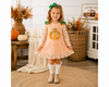 A young girl stands in a cozy, autumn-themed room wearing a peach and white gingham dress with a pumpkin applique on the chest, green leaf shoulder accents, and a green bow in her hair.