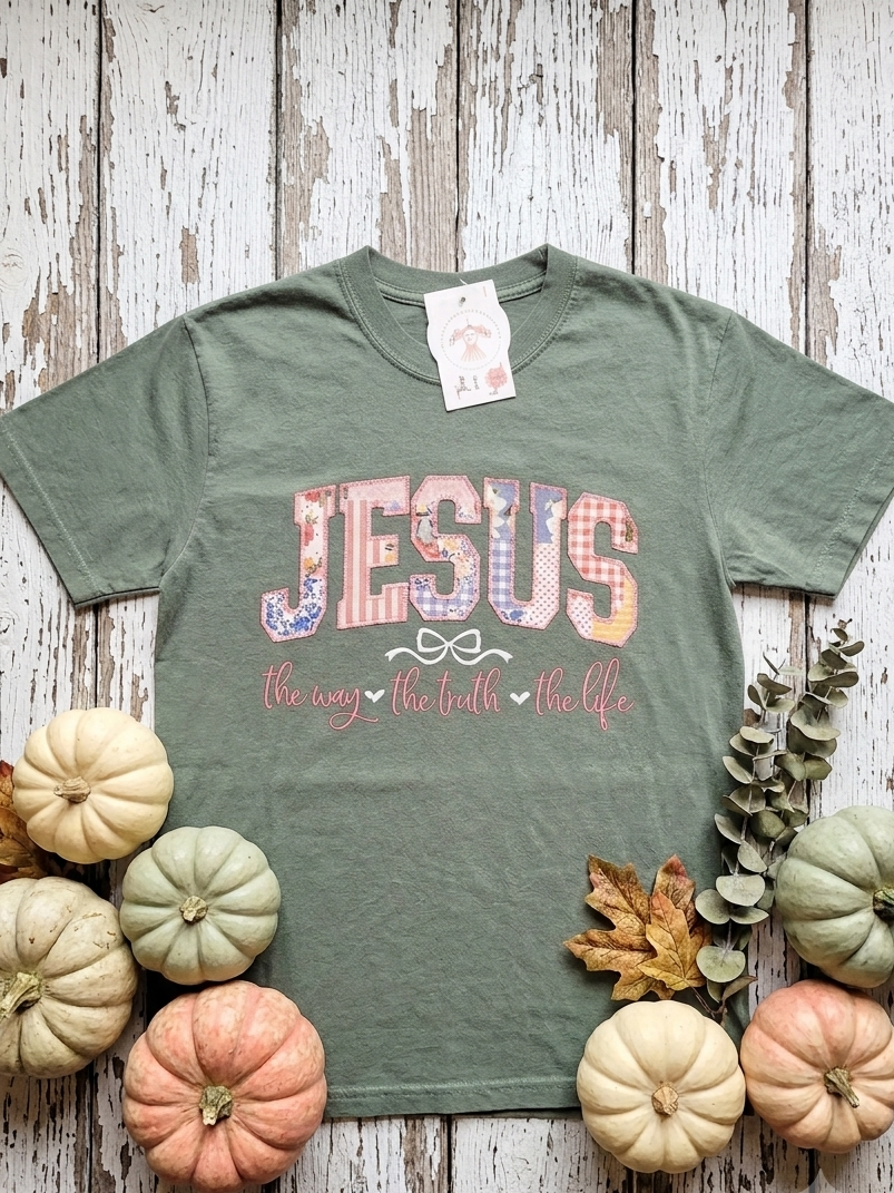 An olive green graphic t-shirt featuring the word 'JESUS' in a patriotic red, white, and blue patterned font, with the phrase 'the way, the truth, the life' in pink script underneath. The shirt is displayed in a flat-lay arrangement on a distressed white wood background, surrounded by small decorative pumpkins and eucalyptus leaves.