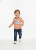 A toddler boy walking forward on a seamless white studio background, wearing a long-sleeve orange and white striped shirt with a blue gingham truck graphic on the chest, paired with dark blue jeans and white sneakers.
