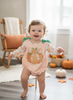 A baby girl wearing a long-sleeve gingham romper with a pumpkin applique and green bow details, standing on a small wooden stand in a cozy, autumn-themed room.