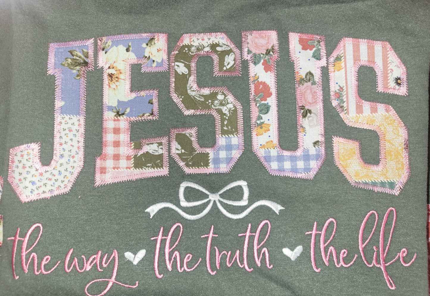 A close-up of a dark green fabric item, likely a sweatshirt or t-shirt, featuring a patchwork-style applique design of the word 'JESUS' in floral and patterned letters, with a white bow graphic and the phrase 'the way the truth the life' in pink script below.