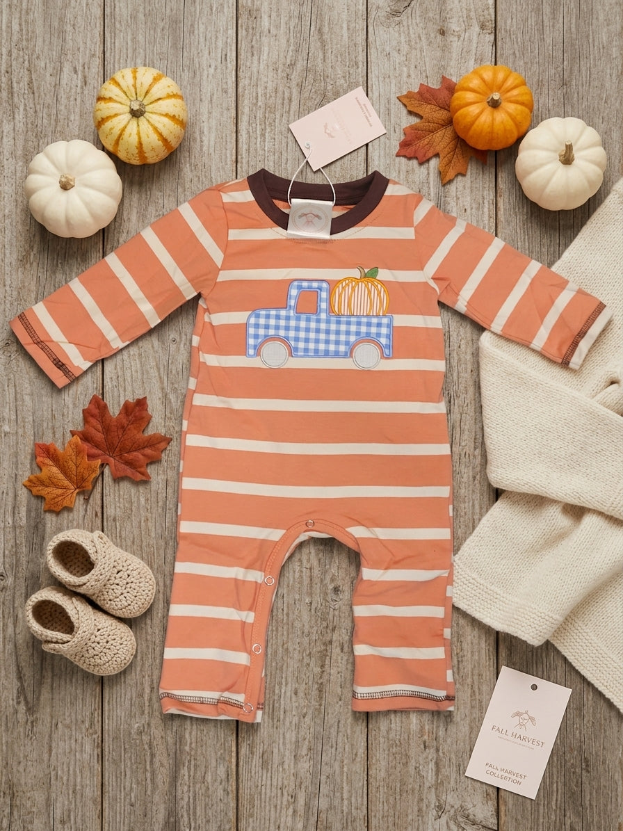 A baby long-sleeve footless romper laid flat on a rustic wood surface, featuring horizontal peach and white stripes and a central applique of a blue gingham truck carrying a small orange pumpkin. The scene is styled with pumpkins, autumn leaves, knit socks, a cream knit blanket, and two tags, one reading Fall Harvest.