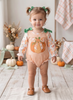 A toddler girl stands in a studio setting wearing a long-sleeve gingham romper in peach and white with a pumpkin graphic on the front, green bow accents at the shoulders, and tan moccasin shoes. The background features a rustic autumn theme with pumpkins, dried flowers, and plaid textiles.