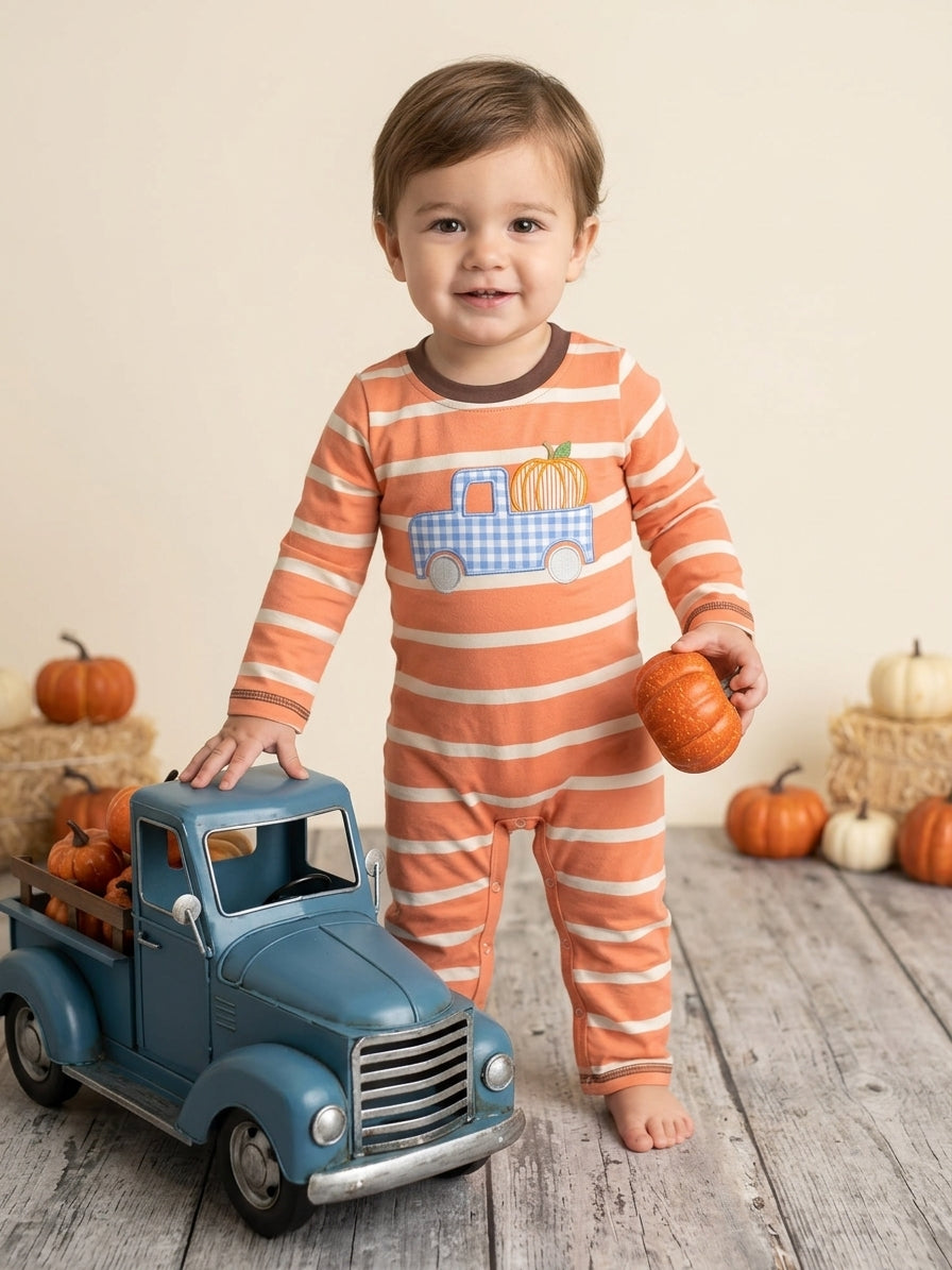 A toddler stands barefoot on a rustic wood floor wearing a peach and white striped one-piece pajama with a blue gingham pickup truck and pumpkin graphic on the chest. The child holds a small orange pumpkin and stands next to a blue toy truck with hay bales and pumpkins in the background.