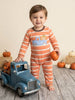A toddler stands barefoot on a rustic wood floor wearing a peach and white striped one-piece pajama with a blue gingham pickup truck and pumpkin graphic on the chest. The child holds a small orange pumpkin and stands next to a blue toy truck with hay bales and pumpkins in the background.