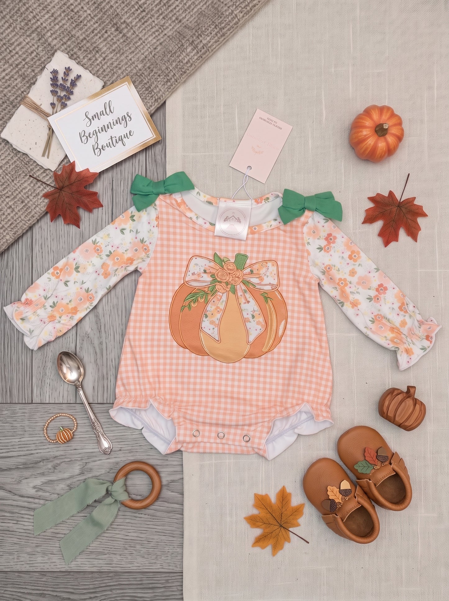 A baby girl's long-sleeve gingham romper in peach and white, decorated with a large orange pumpkin graphic on the front, green bow details at the shoulders, and a white floral print on the sleeves. The romper is styled in a flat lay with autumn-themed props including a small pumpkin, fall leaves, baby shoes, and a card reading 'Small Beginnings Boutique'.