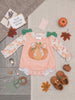 A baby girl's long-sleeve gingham romper in peach and white, decorated with a large orange pumpkin graphic on the front, green bow details at the shoulders, and a white floral print on the sleeves. The romper is styled in a flat lay with autumn-themed props including a small pumpkin, fall leaves, baby shoes, and a card reading 'Small Beginnings Boutique'.
