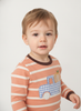 A toddler wearing a peach and white striped long-sleeve shirt with a blue gingham truck and pumpkin applique on the front, photographed against a plain light gray background.
