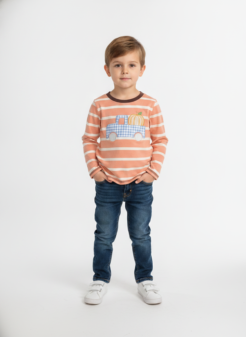 A young boy stands against a plain white studio background wearing a peach and white horizontally striped long-sleeve shirt with a blue and yellow truck graphic on the chest, paired with dark blue jeans and white sneakers.