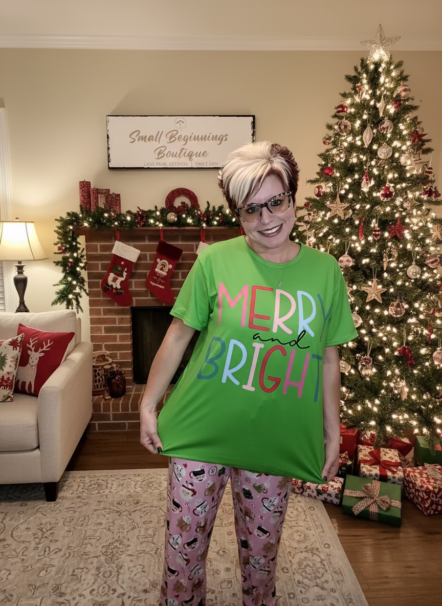 Person wearing a green 'Merry Bright' shirt in front of a wooden wall with 'Small Beginnings Boutique' sign.
