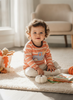 A toddler with curly brown hair sits on a white shag rug in a bright room, wearing a peach and white striped long-sleeve shirt with a blue and white truck graphic. The child is surrounded by autumn-themed toys, including small pumpkins, a basket of fall decorations, and children's books.
