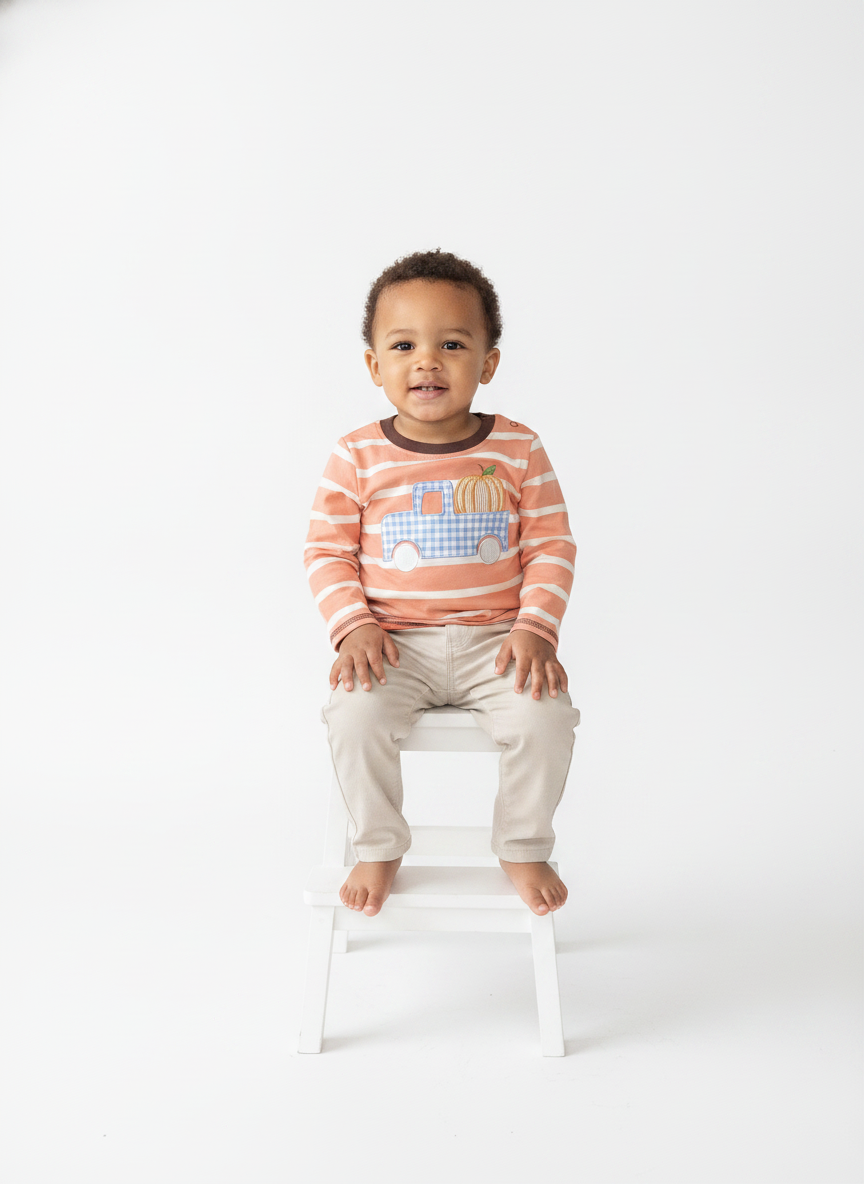 A toddler sitting on a small white stool against a seamless white studio background, wearing a long-sleeve striped shirt in peach and white with a blue and yellow pumpkin truck graphic, paired with light beige pants.