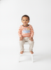 A toddler sitting on a small white stool against a seamless white studio background, wearing a long-sleeve striped shirt in peach and white with a blue and yellow pumpkin truck graphic, paired with light beige pants.