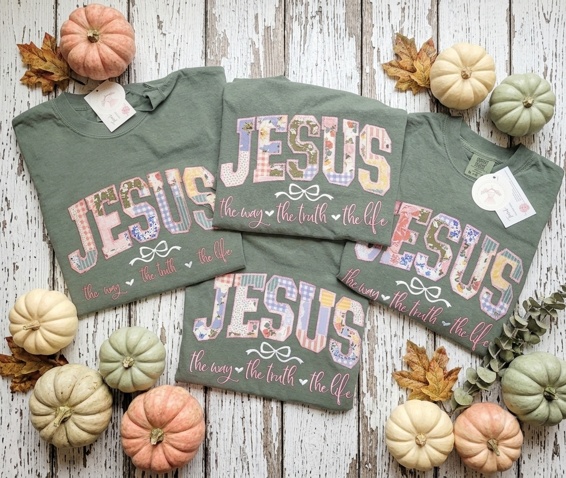 Three olive green graphic t-shirts with 'JESUS the way the truth the life' printed in a floral patchwork pattern, arranged in a flat-lay with small decorative pumpkins and autumn leaves on a white wood background.