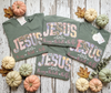 Three olive green graphic t-shirts with 'JESUS the way the truth the life' printed in a floral patchwork pattern, arranged in a flat-lay with small decorative pumpkins and autumn leaves on a white wood background.