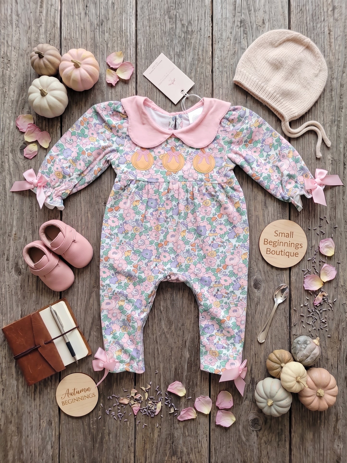 A baby long-sleeve floral romper laid flat on a rustic wood surface, styled with a cream knit bonnet, pink shoes, small pumpkins, petals, and round wooden tags reading 'Small Beginnings Boutique' and 'Autumn Beginnings.' No person or real environment is present, so this is Product/Packaging rather than Lifestyle.
