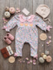A baby long-sleeve floral romper laid flat on a rustic wood surface, styled with a cream knit bonnet, pink shoes, small pumpkins, petals, and round wooden tags reading 'Small Beginnings Boutique' and 'Autumn Beginnings.' No person or real environment is present, so this is Product/Packaging rather than Lifestyle.