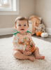 A baby sitting on a white shag rug in a cozy room, wearing a peach and white gingham romper with a pumpkin print and green bow details on the shoulders, holding a small orange pumpkin toy.