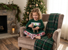 A young girl with curly blonde hair sits cross-legged in a cozy beige armchair, wearing a green Christmas-themed long-sleeve pajama top with a white chest graphic and red-and-white plaid pajama pants. She is holding a small reindeer toy in a decorated living room with a fireplace, a Christmas tree, and a green plaid throw blanket.