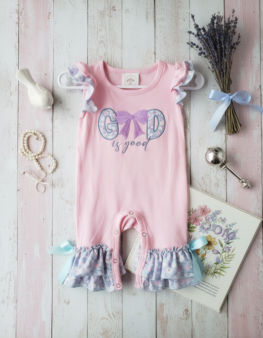Pink baby romper with 'Good is good' text on a wooden surface with decorative items.