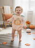 A smiling toddler stands barefoot on a wooden floor in a cozy room, wearing a peach gingham long-sleeve romper with a large pumpkin graphic on the front and green bow details at the shoulders. Pumpkins, autumn leaves, and a rocking chair are visible in the background.