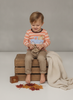 A toddler sits barefoot on a wooden crate wearing a peach and white striped long-sleeve shirt with a blue gingham pumpkin truck graphic, paired with tan pants. A cream knit blanket is draped over the side, and autumn leaves lie on the floor in front.
