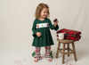 A young girl stands barefoot on a cream studio backdrop wearing a dark green long-sleeve dress with a white chest panel featuring holiday illustrations, red-and-white plaid flared pants, and green bow accents. She holds a pinecone beside a small wooden stool holding a folded red plaid blanket and a white mug.