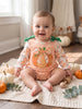 A baby sitting on a cream knit blanket in a cozy room, wearing a long-sleeve romper with a gingham orange and white pattern, a large orange pumpkin graphic on the front, and green bow details on the shoulders.