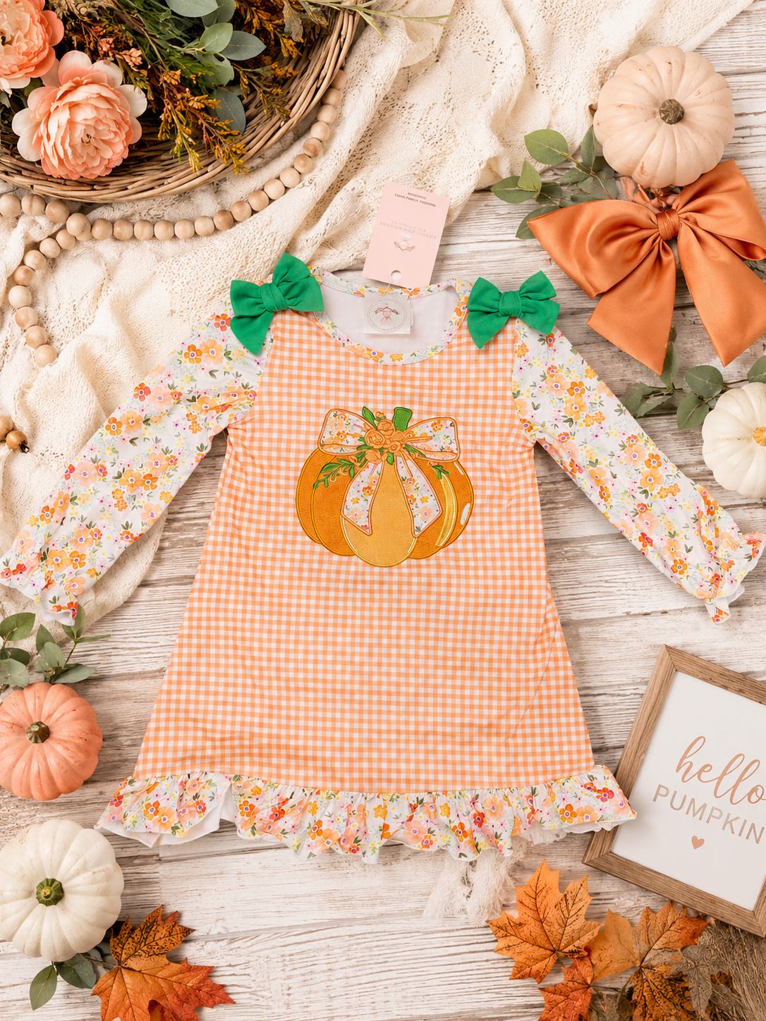 A toddler girl's long-sleeve dress with an orange and white gingham body, floral printed sleeves, green bow details at the shoulders, and a pumpkin applique on the chest. The dress is styled in a flat lay with autumn props including pumpkins, leaves, a lace blanket, and a 'hello pumpkin' sign.