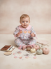 A baby sitting on a beige studio floor wearing a pink floral one-piece jumpsuit with a Peter Pan collar and pumpkin appliques. The scene is styled with small pumpkins, scattered pink petals, a small notebook, and a soft fabric accessory on a warm neutral backdrop.