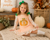 A young girl sitting on a crocheted rug in a cozy fall-themed room, wearing a peach and white gingham dress with a pumpkin applique and green bow accents.