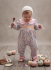 A baby wearing a pink floral long-sleeve footed jumpsuit with a pink Peter Pan collar and a white knit bonnet, standing on a rustic wooden floor surrounded by small pumpkins and a wooden spoon.